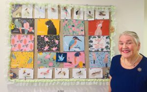Fay, an aged care resident at Regis Kirwan, smiling and holding a crafted item in her hands, surrounded by other residents and team members