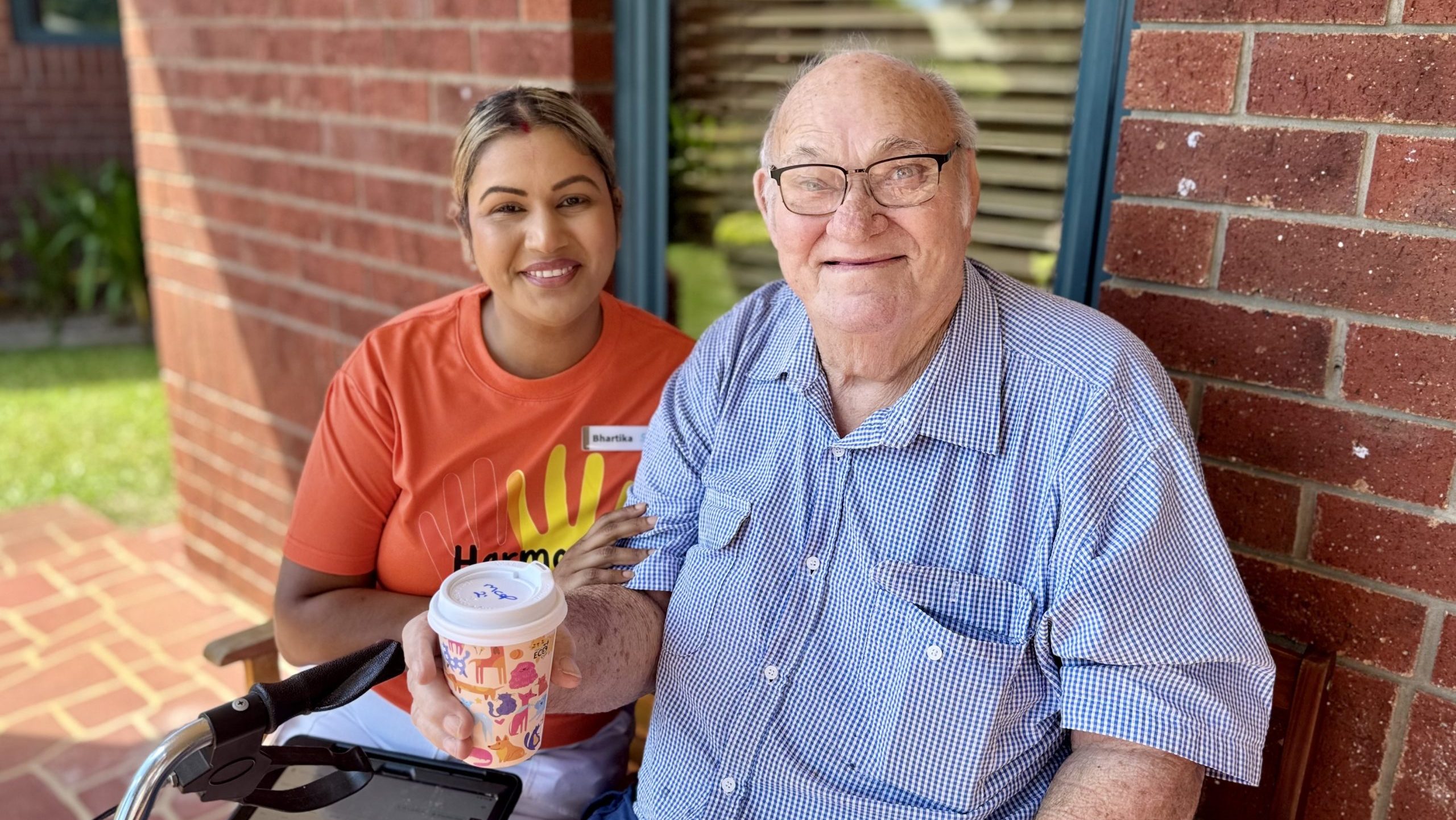 A female aged care employee, wearing an orange t-shirt, sits with a male aged care resident holding a coffee cup
