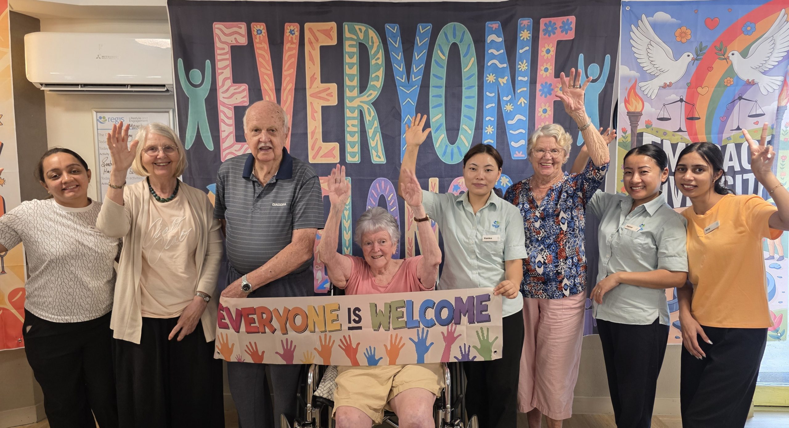 Eight aged care residents and employees standing with their hands up in front of a colourful sign that says "everyone belongs". The person in the middle of the group holds a rainbow sign that says "everyone is welcome"