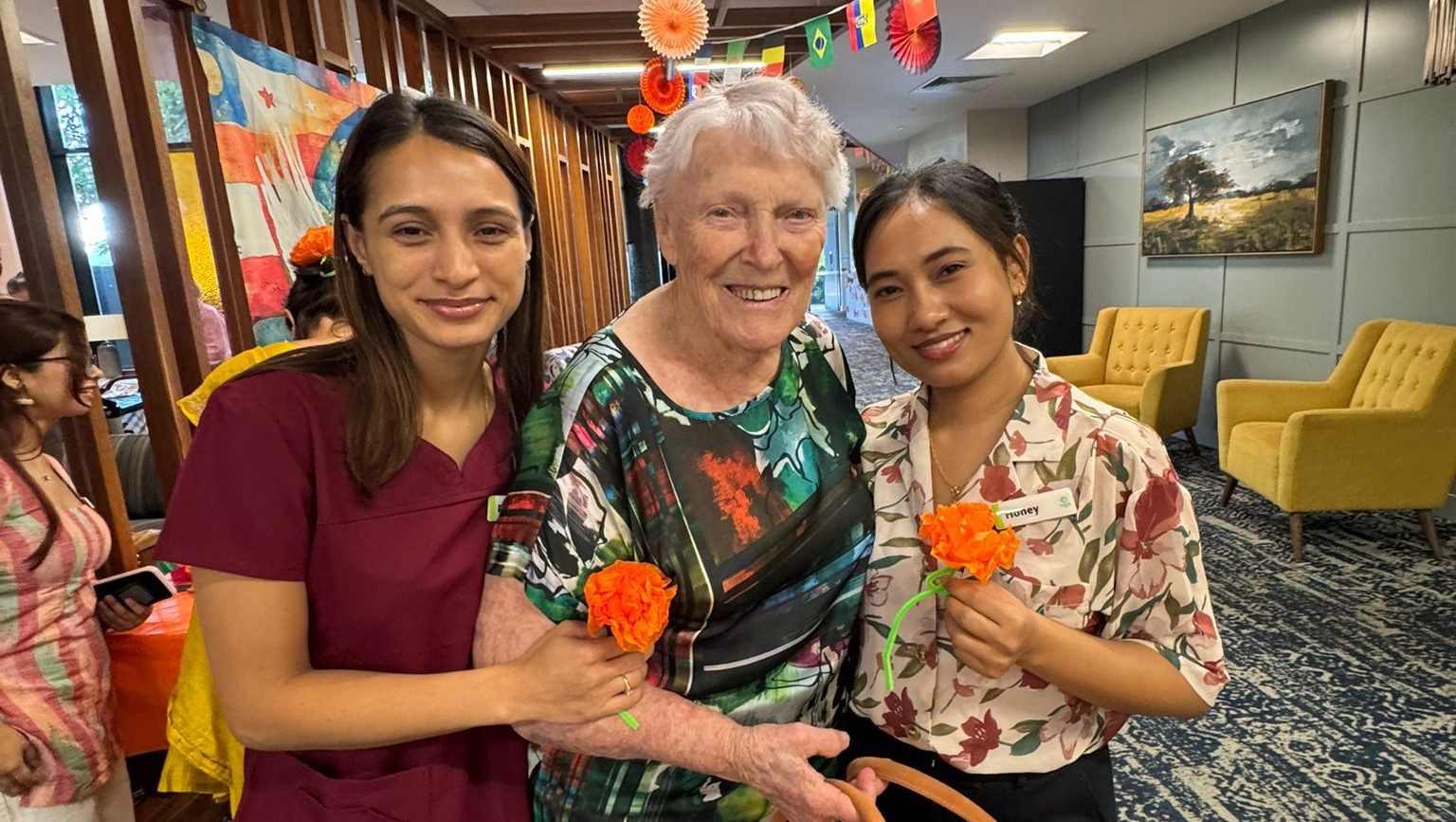 Two female aged care employees stand with a female aged care resident, holding paper orange flowers and smiling
