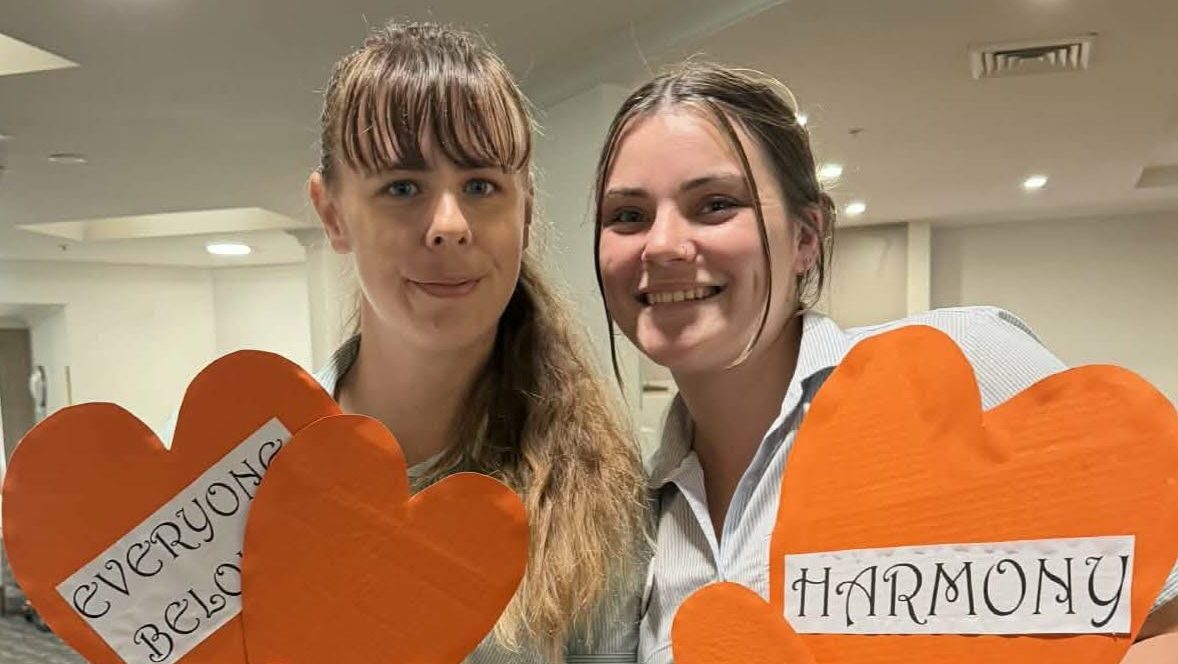 Two female aged care employees smiling, holding paper orange hearts that say, "everyone belongs" and "harmony"