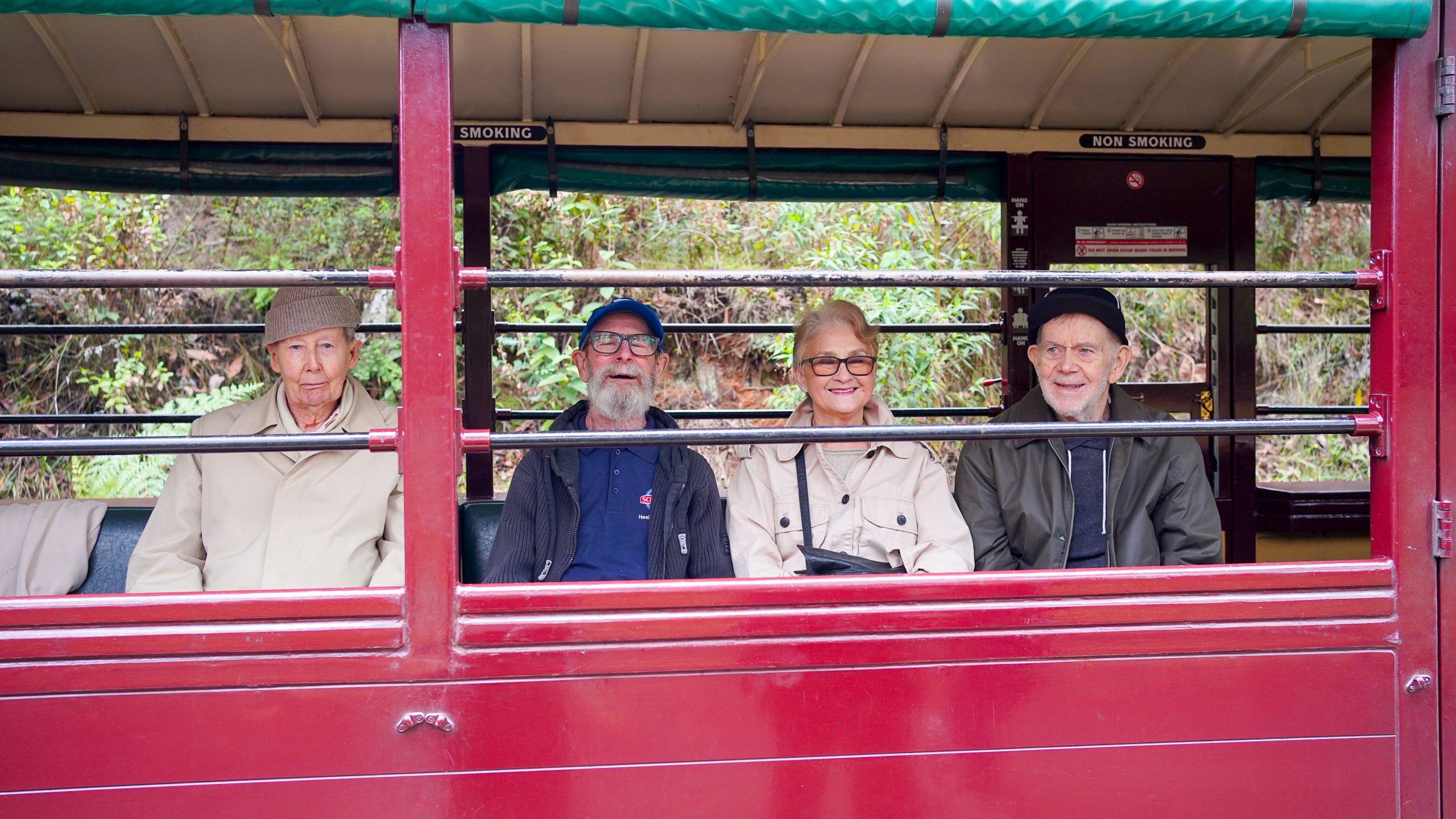Four people sit in a row on an old-fashioned, red, steam train, looking out the open window. There is forestry in the background.