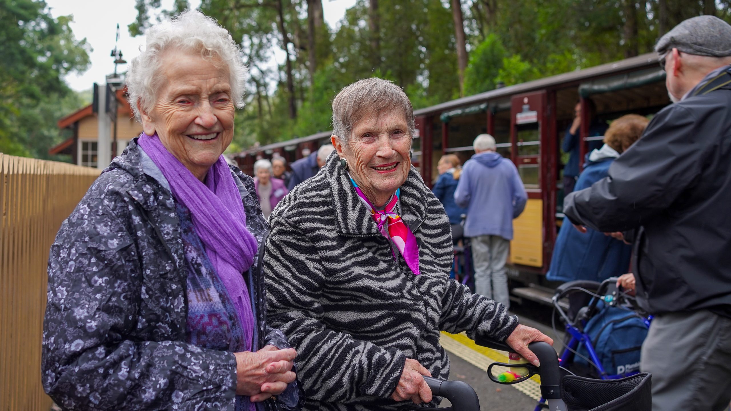 Two women dressed in scarves and jackets stand on a train platform smiling. One is using a walking aid, and the other has her hands clasped together.