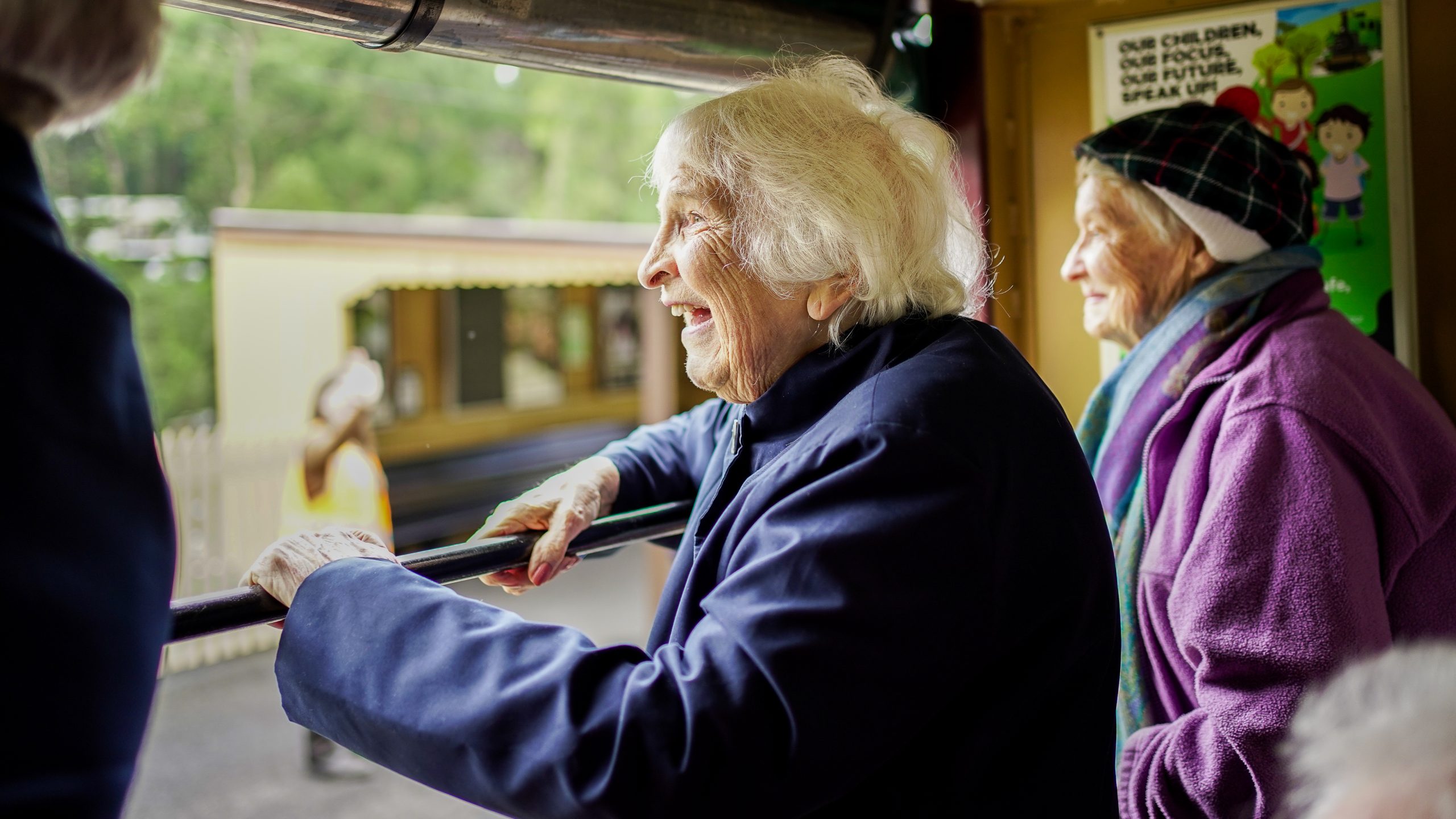 Two elderly women look out a train window to the platform below. They are holding onto the railing and smiling.