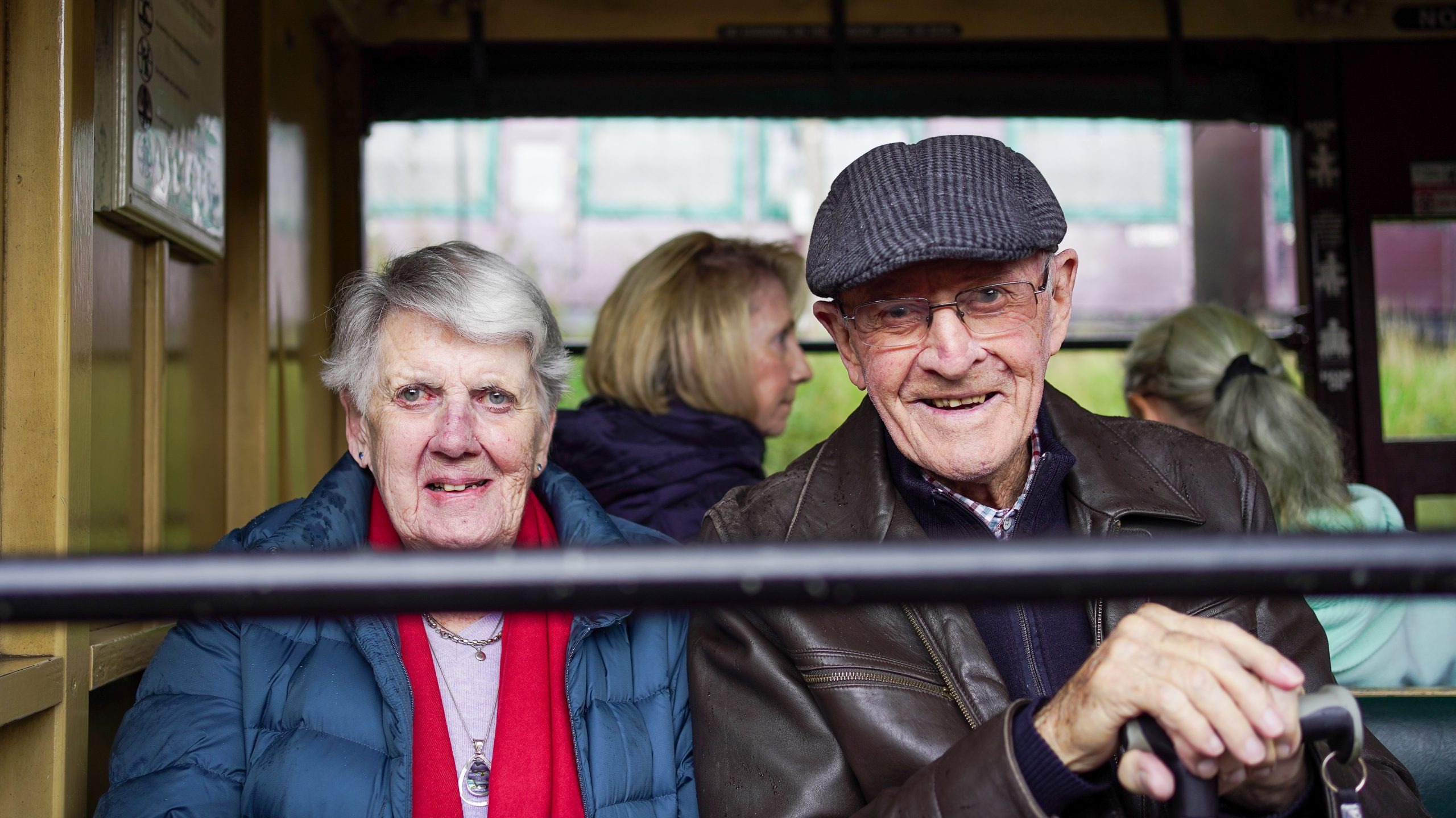 An elderly couple sit smiling from inside an old-fashioned train. They are dressed warmly in scarves and jackets.